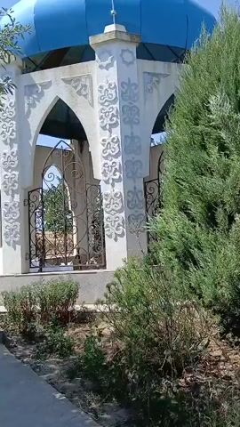 The video shows a tranquil outdoor scene centered around a small, domed structure. The structure is white with decorative carvings on its pillars and features arched openings with ornate metal gates. A bright blue dome, topped with a crescent moon symbol, crowns the structure. The scene is framed by lush green trees and bushes, suggesting a well-maintained garden or park. The sky is clear and blue, indicating pleasant weather. The camera pans slowly, offering different perspectives of the structure and its surroundings. At one point, the view shifts to show people sitting under a covered walkway to the left of the main structure. They appear to be observing something or waiting. The overall atmosphere is peaceful and serene.