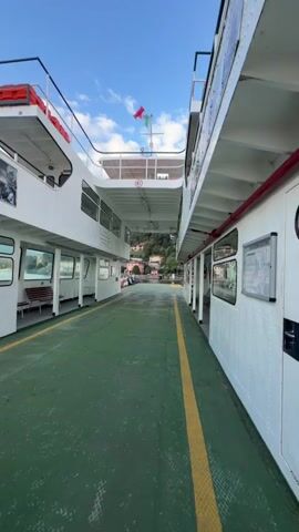 The video shows the interior deck of a ferry. The deck is covered in green material with a yellow line painted down the center. On either side of the deck are the white hulls of the ferry, featuring rows of windows. The windows on the right side appear to look into an enclosed seating area with wooden benches. The windows on the left side also appear to lead to enclosed areas with benches visible. In the distance, the ferry is docked at a port, with buildings and trees visible on a hillside under a blue sky with scattered clouds. A flag is visible on the top deck of the ferry, and a sign with "4.0" is also visible. The scene is static, indicating the ferry is either docked or moving very slowly. There are no people visible in the immediate foreground or moving around the deck.
