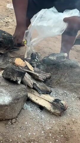 A person, visible from the waist down and arms, is crouched near a small fire pit on sandy ground. The fire pit is formed by large, dark grey stones and contains pieces of wood and ash. The individual wears dark shorts and sandals. They are holding a clear plastic bag in their right hand, directing its open end toward the fire. With their left hand, they hold a lit igniter, applying flame to material being released from the plastic bag onto the existing embers and wood, which produces small flames and smoke. A white bucket is partially visible in the blurred background.