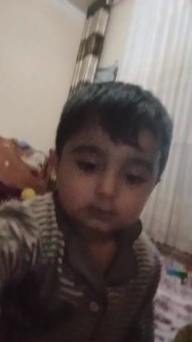 The video features a young child, possibly a toddler or preschooler, facing the camera directly. The child has dark hair and eyes. They are wearing a striped, long-sleeved shirt. The background includes a bed with blankets, a rug with a floral pattern, and a curtained window. The child makes a slight movement with their mouth. The camera appears to be hand-held or resting on a surface, as it is relatively steady. There are no significant actions or interactions beyond the child's slight movement. The lighting appears to be from indoor sources.
