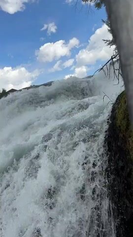 The video shows a powerful waterfall with white water crashing down. The camera is positioned close to the water, offering a dynamic and immersive perspective. In the background, there are trees, buildings, and a cloudy blue sky. The waterfall appears to be quite wide, and the spray from the water creates a misty atmosphere. The overall impression is one of nature's raw power and beauty.