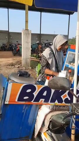 A woman wearing a blue hijab and a patterned scarf stands in front of a blue and orange food cart, looking to her left. Her right hand is extended, touching the cart. In the background, several motorbikes are parked under a covered structure. Two men are visible in the distance, one wearing a yellow shirt and white shorts, the other in a dark shirt. Another person, partially obscured, is on the right side of the frame near the food cart. The ground appears to be dirt or sand. The overall setting seems to be an outdoor market or a roadside stall.