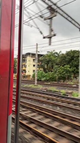The video shows a view from a moving train. The camera is positioned to the right of the frame, capturing the side of the train carriage, which is painted red. The train tracks run parallel to the right side of the frame, with lush green grass and some debris visible along the edges. Beyond the tracks, there are several buildings and dense vegetation, including palm trees. The sky is overcast. As the train moves, different structures come into view, including what appear to be residential buildings, some of which seem to be in disrepair or constructed with simple materials. Electrical poles and wires associated with the railway system are also visible throughout the video. The overall impression is of a landscape that is a mix of urban development and nature, seen from the perspective of a passenger on a train. There are no people clearly visible in the frame.