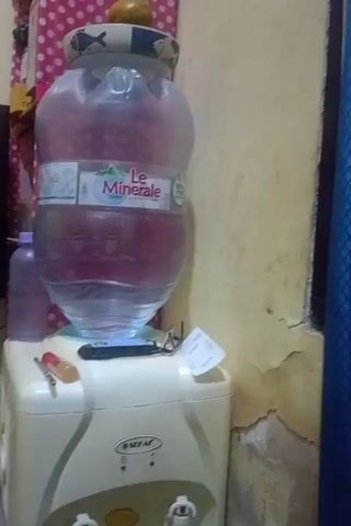 The video shows an indoor scene. A large water dispenser with a bottle of Le Minerale is visible on a white appliance. A small orange fruit is placed on top of the water bottle cap. A tablet computer and other items are on a shelf behind the dispenser. To the left, a television is mounted on the wall. There are also various household items on shelves and surfaces, including a baby bottle sterilizer, a first aid kit, and packaged food. The walls appear to be painted a light color, and the floor is tiled. The lighting suggests it is daytime.