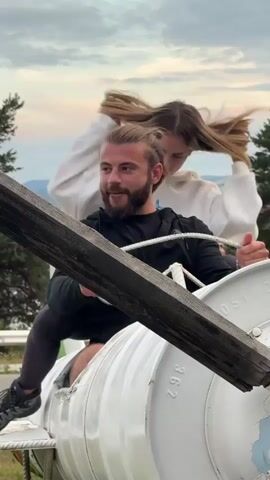 The video shows a man with blonde hair pulled back in a bun and a beard sitting in what appears to be a white cylindrical object. He is wearing a black zip-up hoodie and black leggings. A young woman with blonde hair is sitting behind him, with her arms wrapped around his neck. She is wearing a white hoodie. She leans her head down towards him and touches her face. The man is looking towards the camera and smiling. They are outdoors, with a cloudy sky and trees visible in the background. A wooden beam is positioned diagonally across the foreground, obscuring part of the scene.