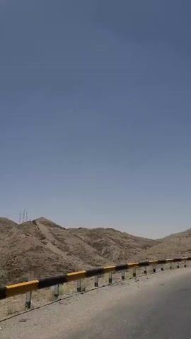 This is a video of a scenic mountain road in Chaman City Tehsil, Pakistan. The time of day appears to be midday with a clear, bright blue sky, indicating sunny and warm weather. The road is paved and curves to the right, with a yellow and black striped guardrail separating it from the rugged, dry, and barren mountainous landscape. The terrain is characterized by rolling hills and rocky outcrops, devoid of significant vegetation, suggesting an arid or semi-arid climate. The colors are predominantly muted earth tones of brown and tan for the mountains, contrasting with the vibrant blue of the sky. The overall mood is one of travel and exploration through a vast, natural environment. There are no people or identifiable objects other than the road, guardrail, and the natural landscape visible in the footage. The camera appears to be mounted on a moving vehicle, likely a car or motorcycle, as indicated by the perspective and the slight motion blur. The video captures the expansive beauty of the mountainous region.