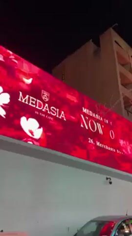 The video shows a brightly lit city street at night. A large LED sign advertising "MEDASIA IN THE CITY NOW OPEN" with red and white floral imagery is visible on a white wall. To the left of the sign are various logos and names, including "TOY ROOM CLUB MALTA," a cartoon bear wearing sunglasses, and the name "PACHA." The street is decorated with strands of lights and palm trees. Buildings are visible in the background.