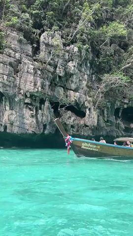The video shows a longtail boat with several passengers and a driver navigating through clear turquoise water near a rocky, vegetation-covered cliff face. The boat is moving from left to right across the frame. The driver, wearing a yellow shirt, is steering the boat using a long oar. The passengers are seated in the boat, some looking out at the scenery. The water is calm and shows ripples from the boat's movement. The cliff is imposing, with several indentations and lush green foliage growing on its surface. The sunlight appears to be diffused, suggesting an overcast or partly cloudy day.