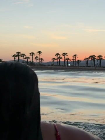 This is a vertical video clip shot from behind a person in the water, facing a beach with palm trees and buildings silhouetted against a vibrant sunset sky. The person has long, dark, wet hair that extends down their back. They are wearing a pink bikini top. The water is calm with gentle ripples, reflecting the soft colors of the sky. The sunset creates a warm, orange and pink hue across the horizon, illuminating the palm trees. The scene evokes a sense of tranquility and relaxation, suggesting a peaceful evening at a tropical or Mediterranean beach. The location context of Castelló de la Plana, Spain, places this scene on the coast of the Mediterranean Sea. There is no visible text in the media.