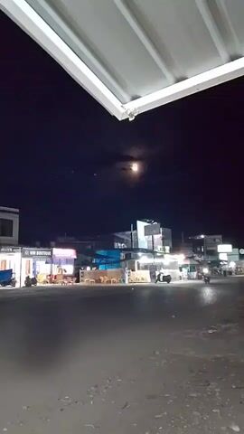 The video captures a nighttime scene in Gampongpaya, Indonesia, under a luminous full moon. The camera is positioned looking upwards, with the underside of a white metal roof or overhang dominating the top portion of the frame, suggesting a sheltered viewing point such as a porch or a covered market stall.

The main subject is the night sky, illuminated by a bright, slightly obscured full moon, with faint clouds drifting across its face. Below the moon, a collection of buildings forms the backdrop. These appear to be shops or small businesses lining a street, their facades lit by various signs and lights. One prominent building has a bright green illuminated sign. Another sign, partially visible, reads "WM BOUTIQUE". Yellow chairs and tables are set up outside some establishments, indicating potential outdoor seating for patrons.

Several motorcycles are visible, parked or in motion, suggesting a street scene in a town or village. One person, wearing a light-colored garment, stands near the tables. The overall atmosphere is calm and quiet, with the nighttime illumination creating a slightly surreal or peaceful ambiance. The paved ground in the foreground suggests a road or a public square. The time of day is clearly night, enhanced by the presence of artificial lights and the moon in the sky. No specific event is discernible, other than the general activity of a village at night.