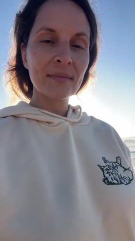 The video shows a close-up of a woman's face. She is wearing a light-colored hooded sweatshirt with a graphic on the left side of her chest. Her hair is dark and pulled back in a ponytail. The background appears to be a beach with the ocean and sky visible. The sun is setting or rising, casting a warm light on her face. The woman is looking directly at the camera and smiling slightly. There is no audio.