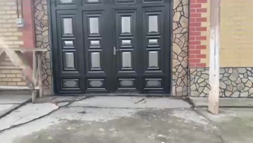 The video shows a stationary, straight-on view of a two-panel metal gate that is black with silver accents. The gate is set into a stone and brick wall that is tan, brown, and red. To the left of the gate is a small wooden structure. To the right is a tan brick wall attached to a tan cement post. The ground is paved and cracked. The lighting is dim.