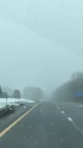 The view from the inside of a car driving on a road during snowy weather is depicted. The road has yellow lines down the middle and white lines on the right edge. There's a patch of snow next to the road. Trees are visible through the fog, and the windshield has snowflakes on it. There's a sign on the left side of the road, but it's blurred by the weather. Another vehicle can be seen far ahead.