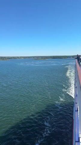 The video shows a wide shot of a body of water under a clear blue sky. The camera is positioned from the deck of a moving vessel, likely a ferry, as indicated by the wake trailing behind and the railing visible on the right side of the frame. The water is a deep blue-green, with gentle ripples on the surface. In the distance, a landmass with trees and buildings can be seen. Several small islands dot the water closer to the shore. A lone sailboat is visible in the middle ground. The overall scene depicts a calm and scenic maritime journey.