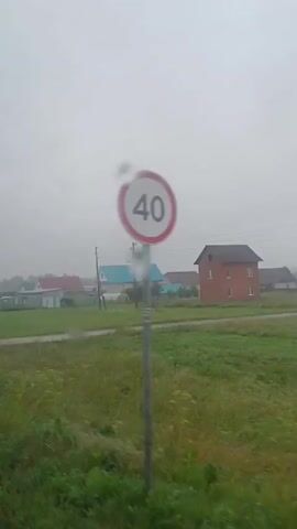 The video shows a rural landscape on a cloudy day. A dirt road runs through a grassy field. In the distance, several houses with red and blue roofs are visible. A power pole stands near the road. A speed bump sign with a speed limit of 20 is posted on a pole. The overall impression is one of a quiet, somewhat overcast, rural setting.