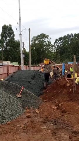 The video shows an outdoor construction site under an overcast sky. A group of construction workers are busy at work. One worker is carrying a yellow bucket on their head, walking towards the center of the site. Another worker, wearing a blue hard hat and a yellow safety vest, follows behind carrying an empty bucket. Several other workers are gathered near a concrete mixer, while others are working in the trenches. In the foreground, there's a mound of reddish-brown soil, scattered with small rocks and construction debris. Two large piles of gravel, one darker than the other, are located to the left of the mound. A shovel rests against the darker gravel pile. In the background, there are trees, a power line, and a fenced-off area.