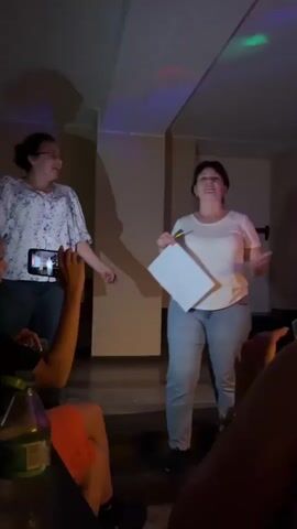 The video shows two women onstage, interacting with a small group of people seated at tables. The woman on the right is wearing a white t-shirt and light-colored jeans and is holding a paper and a pen. The woman on the left is wearing a white top with a blue floral pattern and jeans, and she is holding a smartphone, presumably filming. Other people are sitting at the tables, some with drinks in front of them. The environment is dimly lit, with some colored lights overhead, suggesting an informal event, possibly a party or gathering. There's a wall behind the women onstage, indicating the location is indoors.