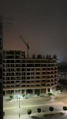 The video shows an elevated view of a street corner at night, centered on a multi-story building under construction. The building is several stories tall with exposed brickwork and open window spaces. A construction crane stands behind it. Streetlights illuminate the road and a small traffic roundabout at the corner. Beyond the building, a city skyline is visible under a dark, overcast sky. The overall light is dim, emphasizing the artificial lighting of the city. There is no visible human activity.
