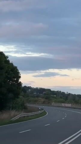 A car is driving on a road. The road is curved and goes uphill. There are trees and houses on the side of the road. The sky is cloudy.