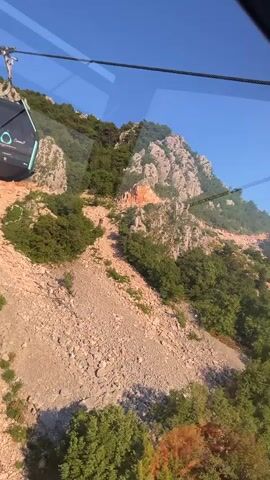 The video captures an outdoor scene from a moving cable car, looking down at a rocky, mountainous landscape covered with green trees and bushes. A large, brown, gravelly area is visible on the hillside. The cable car's cables and a distant support pylon are also seen against a clear blue sky. Another cable car, turquoise and dark-colored, passes by on the left. The camera pans from right to left, revealing more of the lush green and rocky terrain.