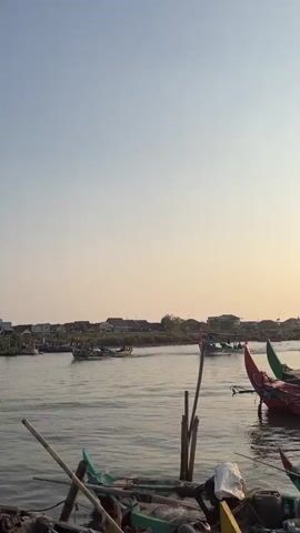 A wide shot captures a body of water with several boats and a shoreline dotted with houses. The sky is clear and a pale yellow hue suggests it is either sunrise or sunset. In the foreground, the edge of a dock is visible, littered with debris including wood and plastic. Several boats are docked here, one green and white, another green. Further out on the water, multiple fishing boats with colorful trim are in motion. Some appear to be moving towards the viewer, while others are moving away. There are people visible in some of the moving boats, though their faces are not discernible. The water is calm with gentle ripples. The houses on the shore are low-rise structures, some with visible rooftops. The overall scene depicts a tranquil waterfront setting.