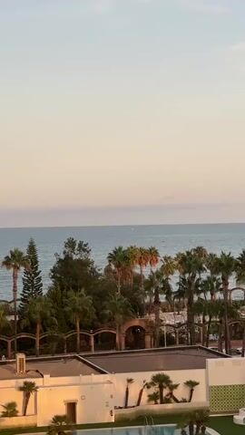 A vertical video captures a serene ocean view with palm trees and resort buildings. The sky is a soft gradient of pale yellow and light blue, suggesting either sunrise or sunset. The ocean is a calm expanse of blue, with a faint horizon line where it meets the sky. Several small boats are visible on the water. In the mid-ground, a lush line of palm trees and other greenery is interspersed with architectural elements, possibly part of a resort or hotel complex, including arched structures. In the foreground, the white facade of a building is illuminated by warm, golden light, hinting at the time of day. A swimming pool, partially visible, adds to the resort ambiance. The overall scene is tranquil and picturesque, conveying a sense of vacation or relaxation. There are no people or discernible actions taking place in the video.