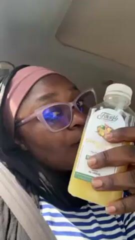 A woman is sitting in the driver’s seat of a vehicle. She is wearing a black head covering, a pink headband, pink glasses, and a black shirt with a blue and white striped design. Her seatbelt is on. She is holding up a bottle of “Four14 Ginger Lemon Bliss” juice, and is looking at the camera. The vehicle appears to be in sunlight.