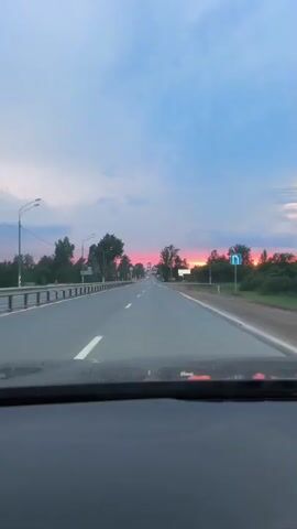 The video shows a first-person perspective from inside a car driving down a paved road. The sky is a blend of blue and pink hues, indicating either sunrise or sunset. Trees and foliage line the sides of the road. There are road signs visible, including what appears to be a directional sign with an arrow. The road has lane markings and a guardrail on the left side. The view is focused on the road ahead, with the dashboard of the car partially visible in the foreground.