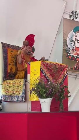 A medium shot captures a man in a red hat and golden robe seated on a raised platform, facing the camera. He holds small instruments in his hands, seemingly playing or using them while speaking into a microphone. He sits behind a table adorned with colorful cloth and floral arrangements in a bucket labeled "Moet & Chandon." The platform is draped in a red cloth. Behind him are cushions with intricate patterns and a framed artwork depicting a spiritual scene. The background appears to be a white tent or structure.