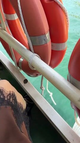 The video, taken from a boat, pans from a person's leg with a black lace skirt past life preservers hanging on the boat's railing and out to the waterfront. The water is turquoise, and the sky is blue. Buildings are visible on the shore, nestled into a steep, green, rocky hillside.