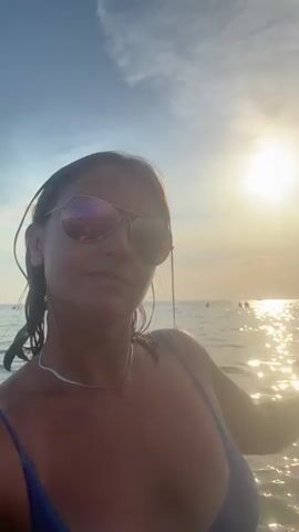 A woman stands in the water, smiling at the camera. She is wearing a blue bikini and sunglasses. The sun is setting behind her, casting a golden glow on the water. There are other people in the water in the background.