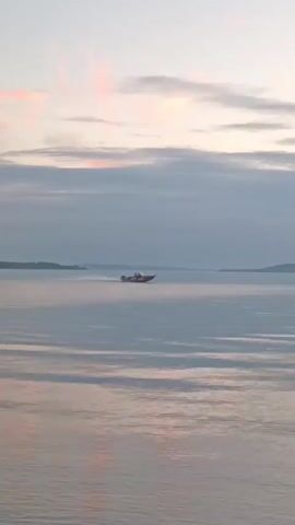 A motorboat speeds across a calm body of water, leaving a wake behind it. The sky above is cloudy with hints of pink and orange as the sun begins to set. A dark, distant shoreline is visible on the horizon.