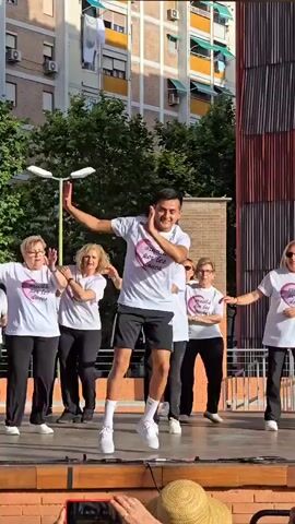 A group of people, mostly elderly women and one younger man, are dancing on an outdoor stage. They are all wearing matching white t-shirts with a pink heart and text on them, and black pants. The younger man is in the foreground, wearing black shorts and white sneakers, and appears to be leading the dance. The women are behind him, mirroring his movements. The stage is wooden and has a brick base. In the background, there are trees, a building with multiple windows and balconies, and a blue sky. The lighting suggests it is daytime. Several people are visible in the lower part of the frame, suggesting an audience, with one person holding up a phone to record.