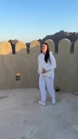 The video shows a woman standing on what appears to be a fort or castle wall, made of light tan-colored stone. She is wearing a white hooded sweatshirt, white pants, and white sneakers. A small white purse hangs across her body. She is holding a phone in her hand. Behind her is a wall with rounded battlements or crenellations, which casts a shadow on the flat surface she stands on. In the background are hills or mountains under a clear blue sky. The camera pans along the wall showing more of the structure, electrical wiring and a white object attached to the wall. The surface of the wall has some weathering and cracks.