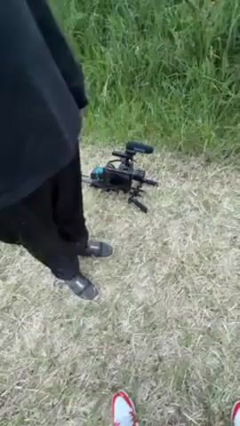 The video begins with a shot of a camera on a tripod on grass. A person is standing near the camera. The camera pans up to a field with tall grass under a cloudy sky. A woman with pink hair is seen walking through the field. She is wearing a white dress and carrying a prop sword. In the background, there is a white barn.