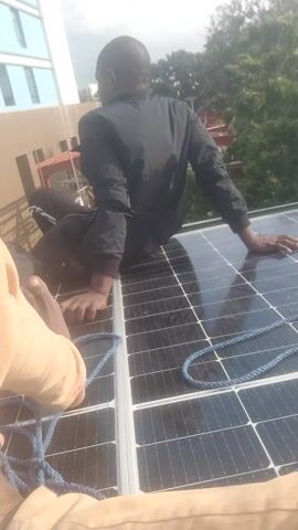 The video shows two individuals working on solar panels. One person, wearing a black jacket, is positioned on top of a solar panel, with their right hand touching the surface. Their left hand appears to be gripping something below the panel. A blue rope is visible on the solar panel. The second person's arms and hands, wearing light-colored sleeves, are in the foreground, also near the solar panels and the rope. The background reveals trees and a building with a modern architectural design. The lighting suggests it is daytime. The overall impression is that of a solar panel installation or maintenance task being performed on a rooftop or elevated surface.