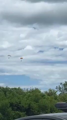 Here is a concise description of the video:

The video captures a lone parachutist descending against a cloudy sky. The parachute is predominantly yellow with black accents. The view is partially obstructed by a light pole and treetops. Several cars are visible in the foreground, suggesting the recording location is a parking lot or similar area.