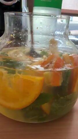 A close-up, slightly shaky, eye-level shot shows a glass pitcher filled with a clear liquid and various fruit slices and green leaves. The pitcher is sitting on a wooden surface, likely a table. The fruit appears to be oranges and lemons, cut into thin slices. Green leaves, possibly mint, are also visible throughout the pitcher. The liquid is somewhat cloudy, and condensation is visible on the outside of the pitcher. In the background, a green and white bottle of Sprite is partially visible. A hand holding a spoon is seen stirring the contents of the pitcher. The lighting is bright and appears to be natural daylight.