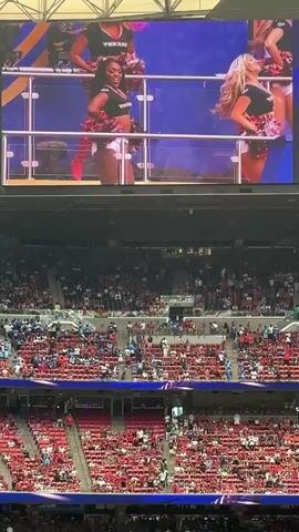 The video shows a wide shot of a stadium with a large screen displaying a performance of cheerleaders. The cheerleaders are dressed in red and white outfits, and they are dancing and holding pom-poms. The screen also shows the FIFA World Cup 2026 logo. Below the screen, the stadium is filled with spectators wearing red clothing. The environment appears to be an indoor stadium with tiered seating.
