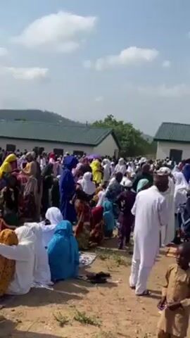 The video shows a large group of people, including adults and children, gathered outdoors on a sunny day. Many are wearing traditional Muslim clothing, including headscarves and robes, in various colors. Some people are sitting on the ground, while others are standing and walking around. There are a few low buildings with green roofs visible in the background, as well as trees and a distant hill. The ground appears to be a dirt or grassy area. The overall scene suggests a gathering for prayer or a religious event.