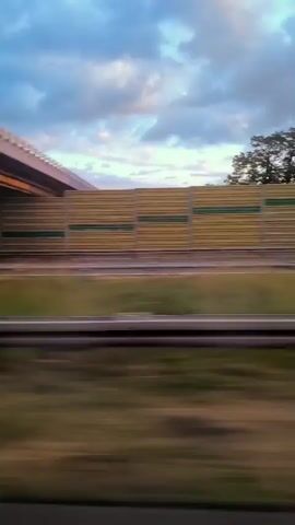 This is a POV shot from a moving vehicle, likely a car or bus, traveling along a road. The view is primarily out the side window. In the foreground, the landscape is blurred due to motion, showing green grass and some darker vegetation. A highway or main road runs horizontally across the middle ground, also appearing slightly blurred. Beyond the road, there is a long, horizontal sound barrier, a light beige structure with horizontal green accents. The sky above is filled with clouds, and the sun is setting, casting a warm orange and yellow glow, especially on the right side of the frame. The overall impression is of a sunset viewed from a roadside. There are reflections visible on the window, including what appears to be the interior of the vehicle. No people are visible.