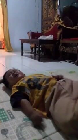 The video shows a young child lying on their back on a mat. The child is wearing a yellow t-shirt with black sleeves and a graphic on the front. The child is crying and appears to be upset or in distress. The child's hands are near their face. In the background, there is furniture, including a wooden table with items on it, and a red chair. There is also a curtain on the left side of the frame. The floor appears to be tiled. The lighting suggests an indoor environment.