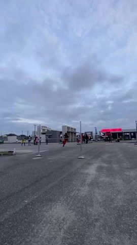 The video shows a roller skater in red pants and a red helmet performing a jump. The skater is positioned between two poles. After a brief hesitation, the skater lunges forward, performs a jump, and lands. The skater then quickly slows down and looks toward the camera. In the background, some people and buildings can be seen, as well as other roller skaters and a cloudy sky.