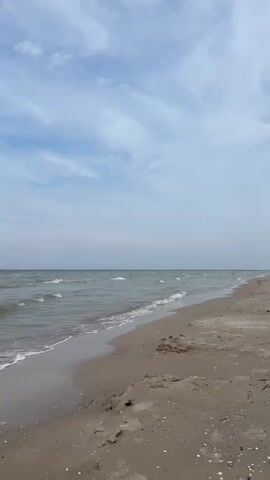 The video presents a tranquil, wide-angle shot of a sandy beach and the sea on a cloudy day in Liepaja, Latvia. Gentle waves consistently roll onto the light brown shore, creating foamy white lines as they spread and then recede. The water appears a muted greenish-grey, extending to a hazy horizon where it meets the sky. The sky is partly cloudy with soft, diffuse white and light blue clouds, indicating daytime. The sandy beach in the foreground is largely undisturbed, showing only natural undulations and traces of the tide. There are no people, objects, or visible text in the frame, emphasizing the natural and untouched beauty of the coastal landscape. The scene conveys a sense of calm and natural serenity.