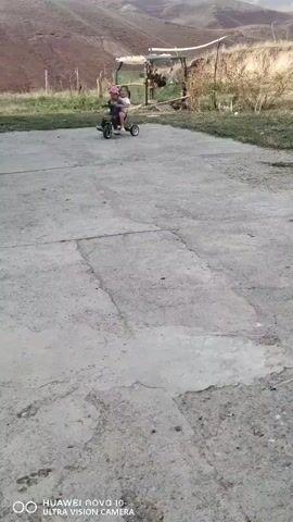 A child wearing a pink hat rides a black tricycle with another child behind them. The tricycle is on a paved surface in front of a dirt hill. There is a small structure with a white roof behind them.