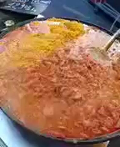 A large metal pan filled with a vibrant, multi-colored dish is shown. The dish appears to be a stew or curry, with various textures and shades of orange, yellow, and white. A spoon is visible in the pan, resting in the mixture. The background is slightly blurred, but hints of a dark surface and possibly some signs or labels can be seen. The overall impression is of a culinary creation, likely being prepared or served.