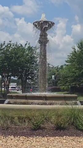 This vertical video showcases a tiered water fountain in an outdoor setting. The fountain is the central subject, with water cascading down its ornate structure and splashing into the circular basin below. Surrounding the fountain is a landscaped area featuring mulch, decorative stones, and various grasses and small plants. In the background, lush green trees line the perimeter, and a few buildings are partially visible, suggesting a developed area, possibly a commercial or residential complex. Beyond the trees, a parking lot is visible with several vehicles, including a white SUV. The sky above is blue with scattered white clouds, indicating a bright, sunny day, likely midday. The overall impression is one of a well-maintained public space, perhaps a town square or a common area in a community, offering a serene and refreshing ambiance. There is no visible text in the video.