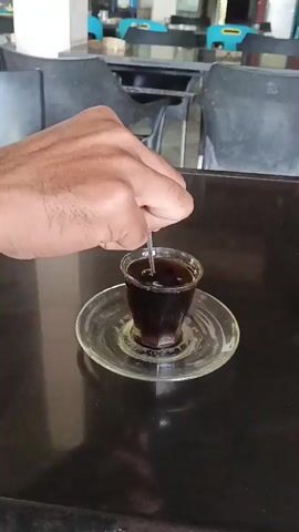 A hand is seen stirring a dark liquid in a small clear glass cup placed on a clear glass saucer. The glass is sitting on a dark reflective surface, possibly a table. The background is blurred but shows chairs and some colorful objects in what appears to be an indoor setting like a cafe or restaurant. The person's hand is positioned from the top right of the frame, and they are holding a utensil, likely a spoon or stirrer, which is submerged in the liquid. The liquid appears to be coffee or a similar dark beverage.