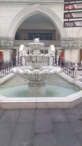 The video shows a three-tiered marble fountain with water flowing from the top tier into the lower tiers and then into the basin. The fountain is set against a backdrop of a white building with arched entrances and ornate columns. There are black metal railings on either side of a set of stairs leading up to the building. A few signs are visible on the right side of the building, one of which indicates "ECZANE PHARMACY". The ground in front of the fountain is paved with large gray rectangular tiles. The water in the fountain appears to be clear and slightly turquoise. The overall scene suggests an outdoor public space, possibly a plaza or courtyard in front of a commercial establishment.
