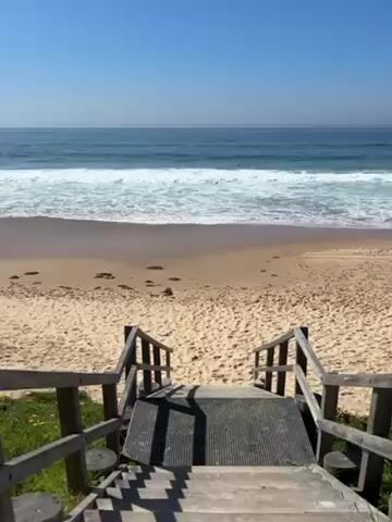 The video shows a view from the top of wooden stairs leading down to a sandy beach. The ocean is visible in the background, with waves breaking on the shore and a few people surfing. The sky is clear and blue. There is some greenery on either side of the stairs.